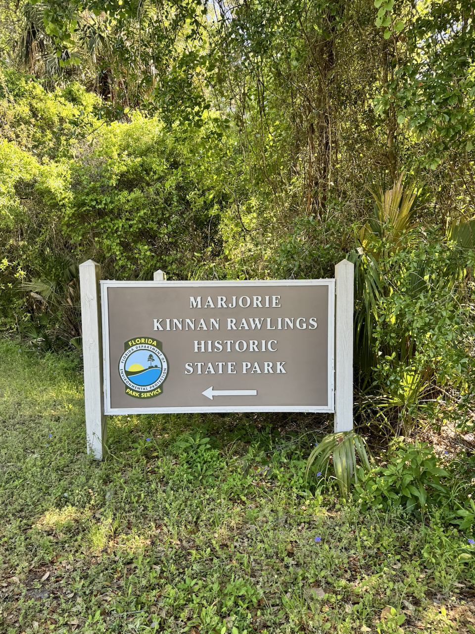 Brown state park entrance sign reading 'Marjorie Kinnan Rawlings Historic State Park' with the Florida Department of Environmental Protection Park Service seal, surrounded by subtropical vegetation.