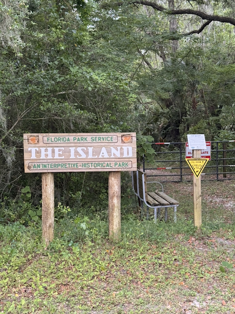 Wooden sign reading Florida Park Service, The Island, An Interpretive-Historical Park, standing at a trail entrance in dense forest with a bench and gate behind it.